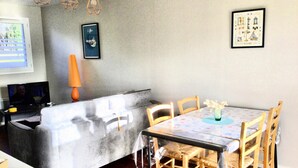 Un comedor con una mesa redonda, sillas y un florero. Hay un televisor sobre un mueble bajo y un cuadro en la pared.