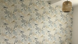 Un dormitorio con papel tapiz floral, una cama con manta azul, un cojín amarillo y una mesita de noche con una lámpara.