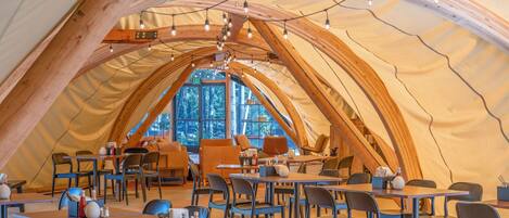 A spacious tent with multiple tables and chairs, a wooden ceiling, and a view of the forest through large windows.