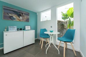 A modern room with teal walls, a white cabinet, two blue chairs, a small white table, and a large window with a view of greenery. - Alsol Los Caribes 2 (San Bartolomé de Tirajana)