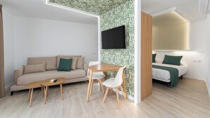 A modern hotel room with a sofa, a dining table with chairs, a TV mounted on the wall, and a bed with green bedding. - Alsol Los Caribes 2 (San Bartolomé de Tirajana)