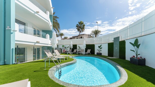A swimming pool with a surrounding grassy area, lounge chairs, and a modern building with balconies. - Alsol Los Caribes 2 (San Bartolomé de Tirajana)