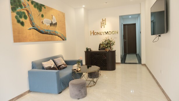 A modern hotel lobby with a blue sofa, a round ottoman, a coffee table, and a painting of birds on a branch. The reception desk is visible with a sign reading "Honeymoon." - HONEYMOON HOTEL 2 (Hanoi)