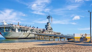 A large naval ship docked at a pier with a sandy beach and a clear blue sky in the background. - Lighthouse at the beach (Corpus Christi)