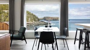 A dining area with a table and chairs, large windows with curtains, and a view of the ocean and hills. - Cliffs Edge Retreat (Tors Cove)