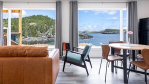 A modern living room with a view of a coastal landscape, featuring a sofa, chairs, and a dining table. - Cliffs Edge Retreat (Tors Cove)