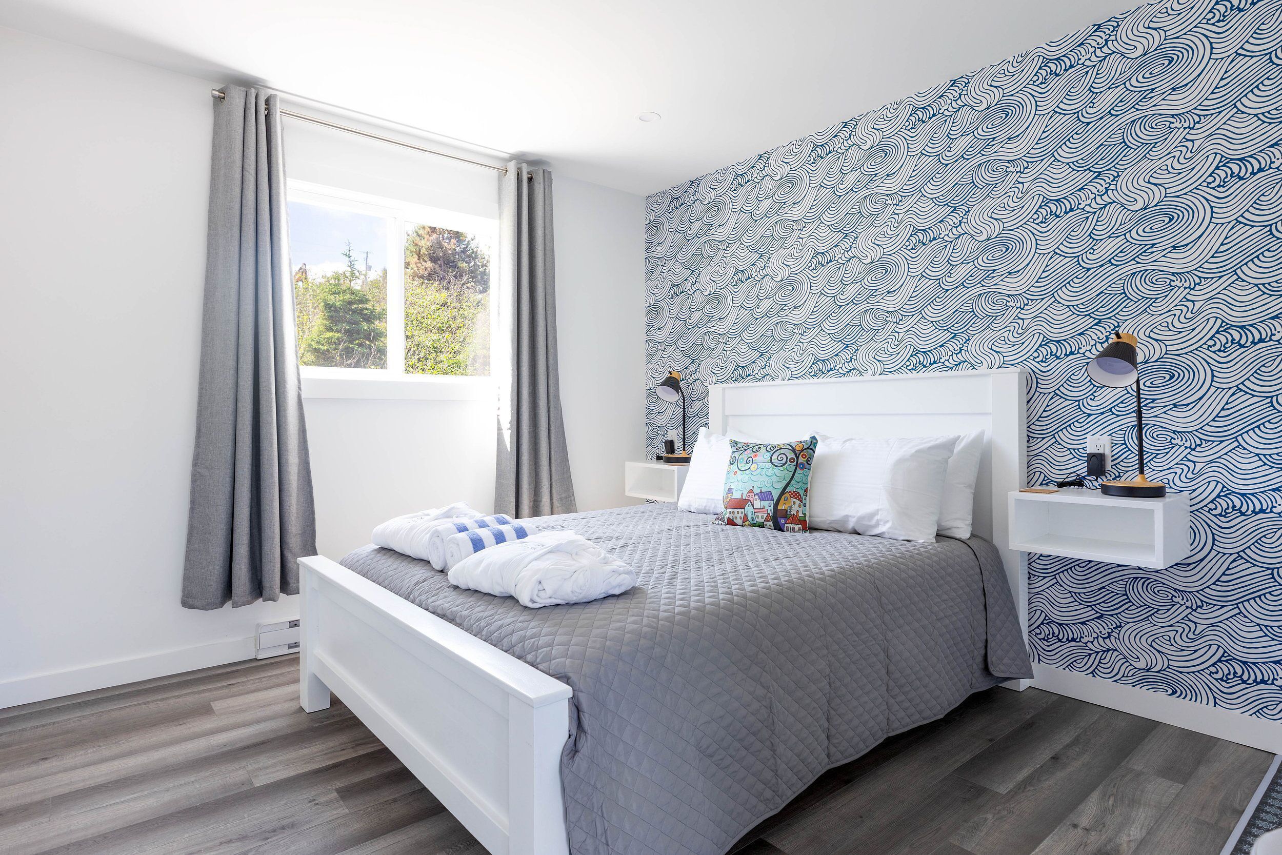 A modern bedroom with a grey quilted bedspread, a white headboard, and a patterned wallpaper.