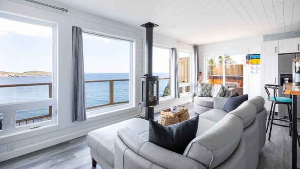 A modern living room with a large window overlooking the sea, a fireplace, and a cozy seating area. - Cliffs Edge Retreat (Tors Cove)
