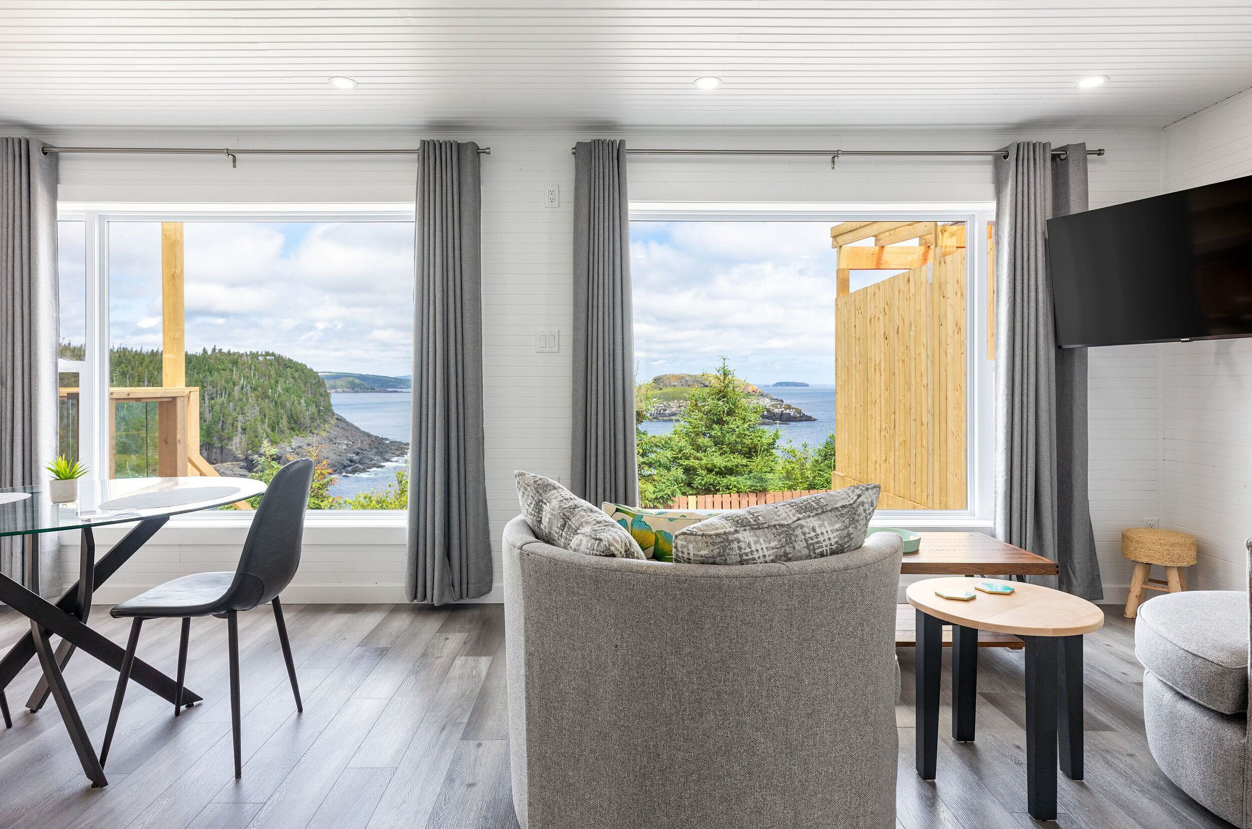 A modern living room with a large window, a flat-screen TV, and a dining area with a glass table and chairs.