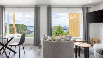 A modern living room with a large window, a flat-screen TV, and a dining area with a glass table and chairs.