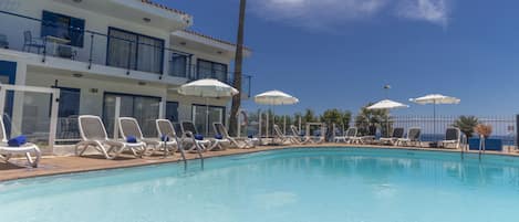 A pool area with lounge chairs and umbrellas, a building with balconies, and a clear blue sky.