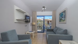 A modern living room with two grey sofas, a small white table, a mounted TV, and a balcony with a view of buildings. - Alsol Los Caribes 1 (San Bartolomé de Tirajana)