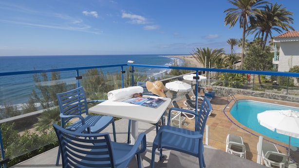A balcony with a pool, chairs, and a table overlooking the ocean. - Alsol Los Caribes 1 (San Bartolomé de Tirajana)