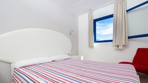 A bed with a colorful striped bedspread, a white headboard, a window with blue trim, and a red chair. - Alsol Los Caribes 1 (San Bartolomé de Tirajana)