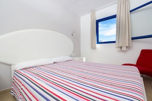 A bed with a colorful striped bedspread, a white headboard, a window with blue trim, and a red chair. - Alsol Los Caribes 1 (San Bartolomé de Tirajana)