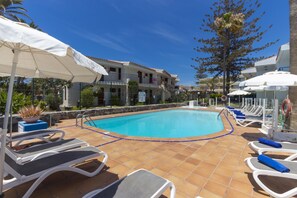 A swimming pool with lounge chairs and umbrellas, surrounded by buildings and palm trees. - Alsol Los Caribes 1 (San Bartolomé de Tirajana)
