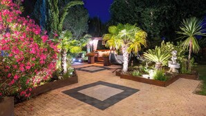 Uma área de jardim pavimentada com espaço para sentar, rodeada por diversas plantas e uma estátua.