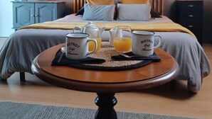 Uma cama bem arrumada com travesseiros, uma mesinha de cabeceira com duas chávenas e um bule, e uma parede com ripas de madeira verticais.