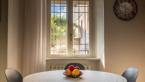 Zona de menjador amb taula rodona, dues cadires, un plat de fruita, finestra amb cortines i un rellotge de paret.