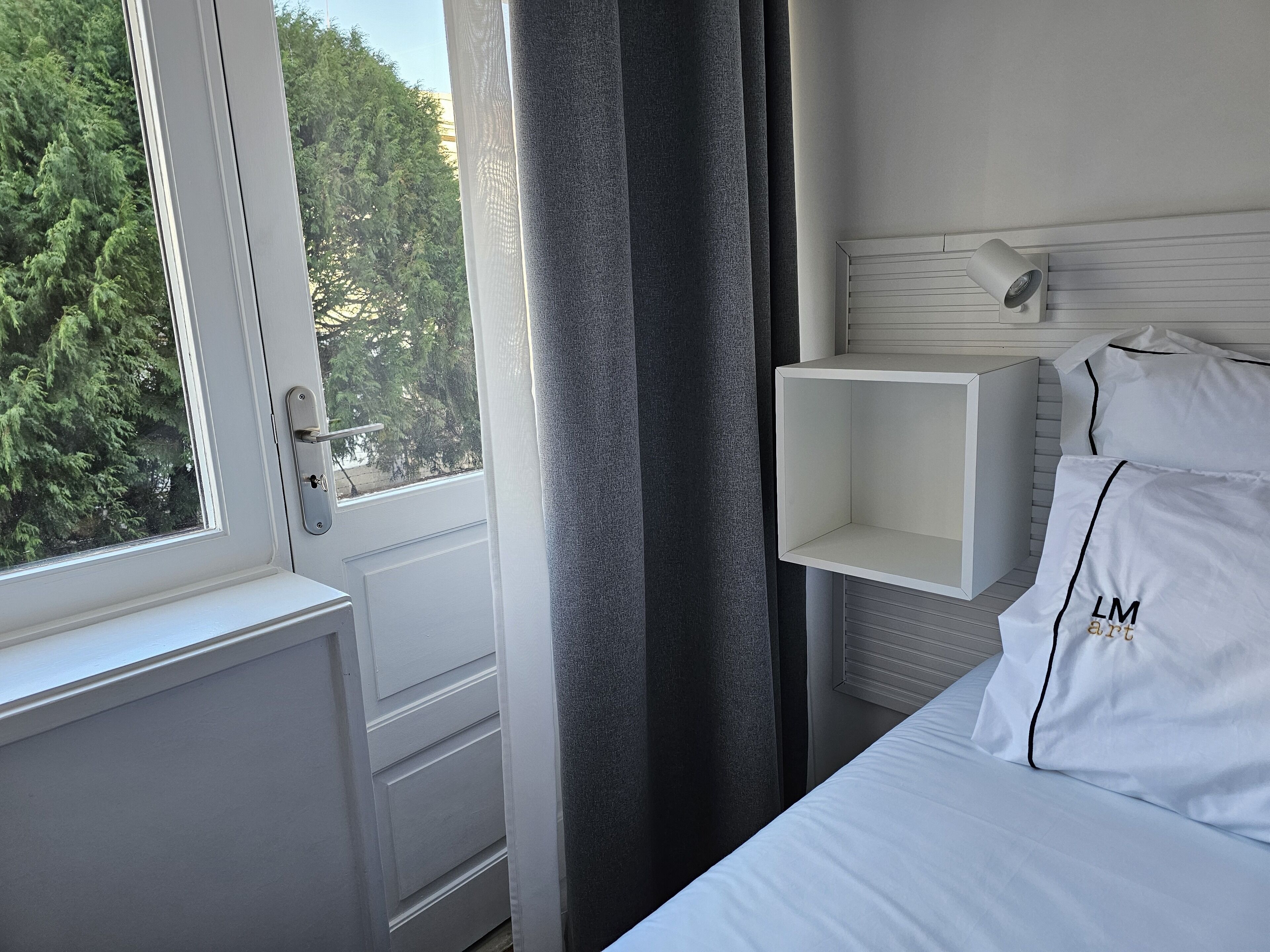 Habitación de hotel con cama, ventana con vista a árboles, mesita de noche blanca y lámpara fijada a la pared.