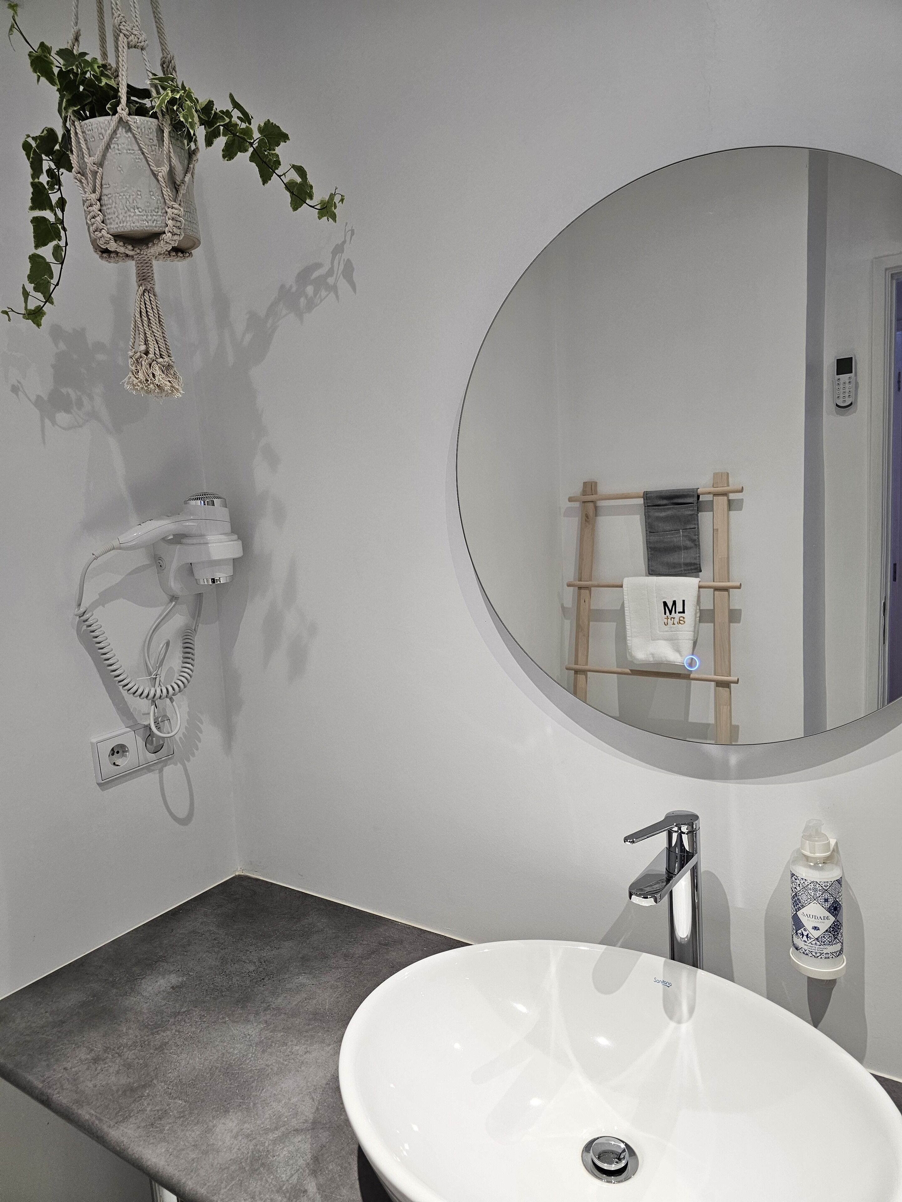 Une salle de bain avec un miroir rond, un lavabo blanc, un sèche-cheveux, un porte-serviettes et une plante.