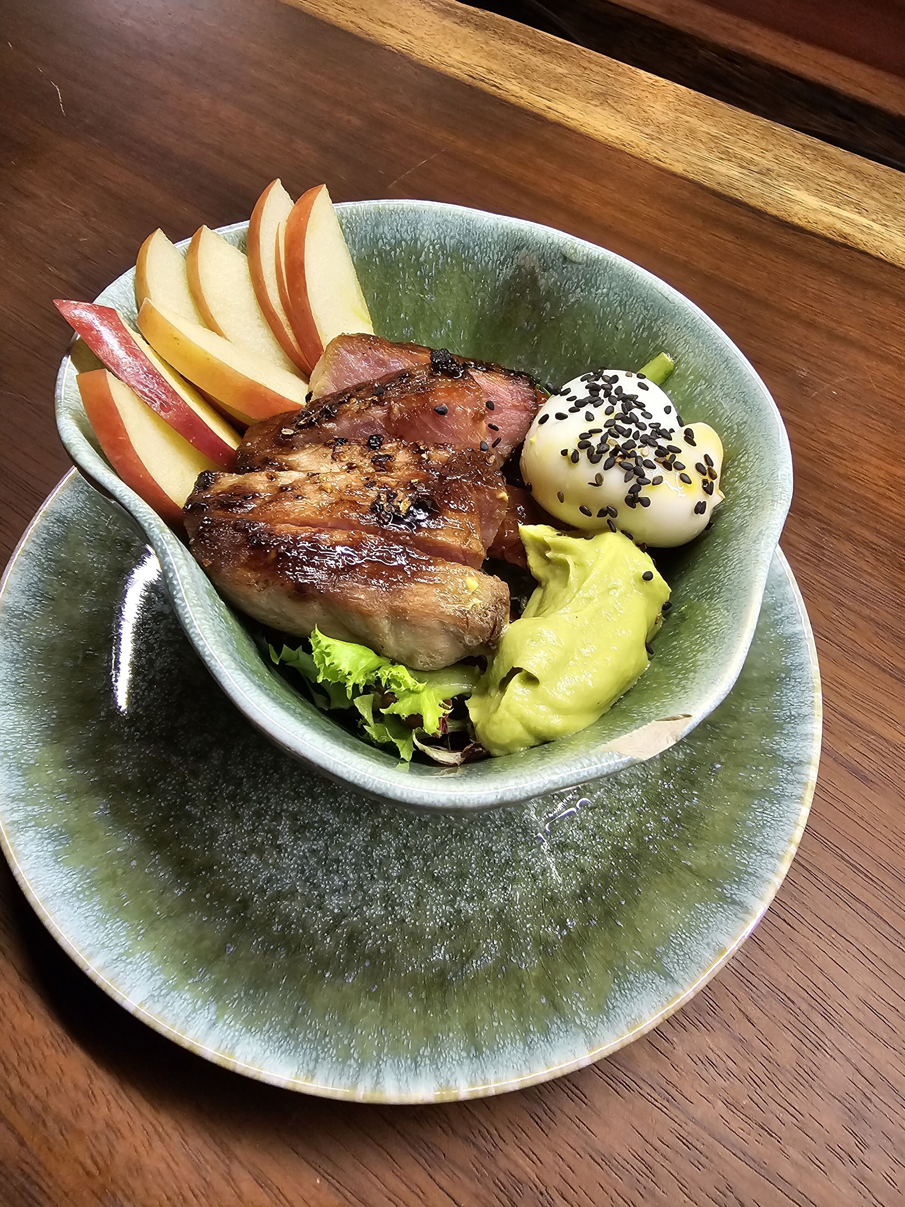 Uma tigela com comida, incluindo fatias de maçã, um pedaço de carne e um bolinho, sobre um prato verde.