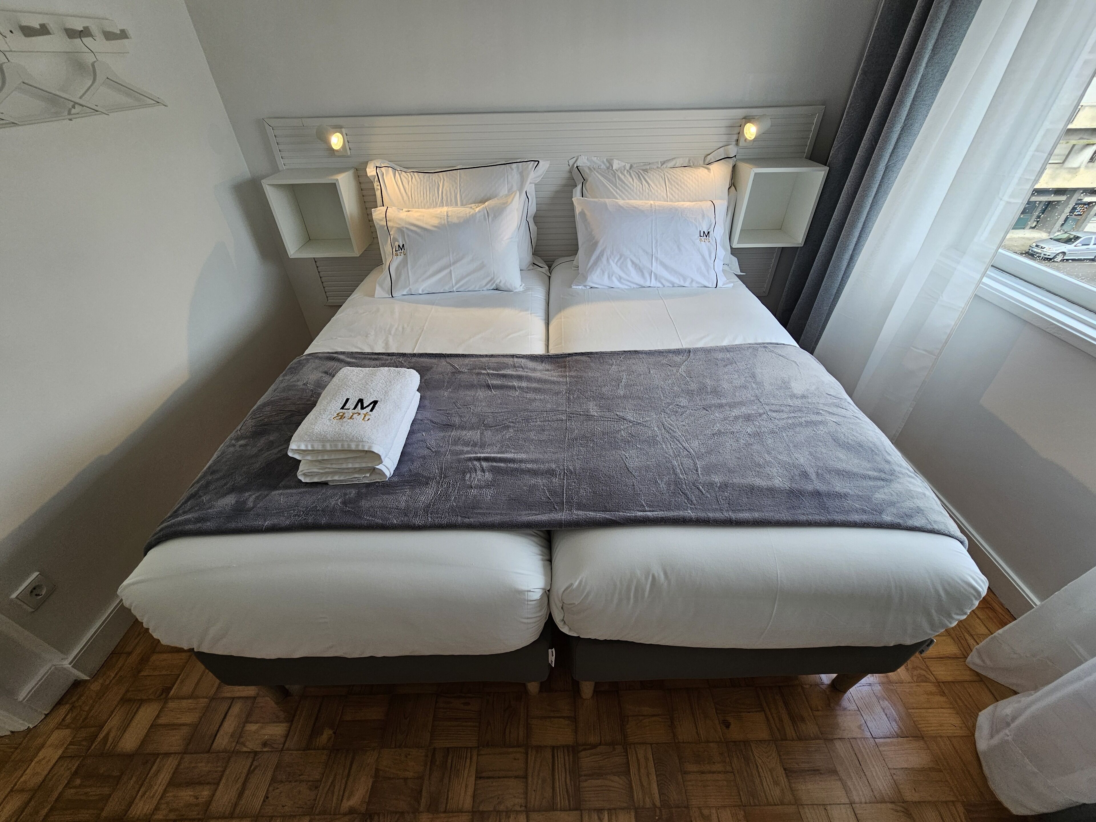 Ein Doppelbett mit weißen Laken, einer grauen Decke und einem Handtuch mit dem Aufdruck 'LM art'. Zwei Nachttische mit Lampen, ein Fenster mit Vorhängen und ein Holzboden.
