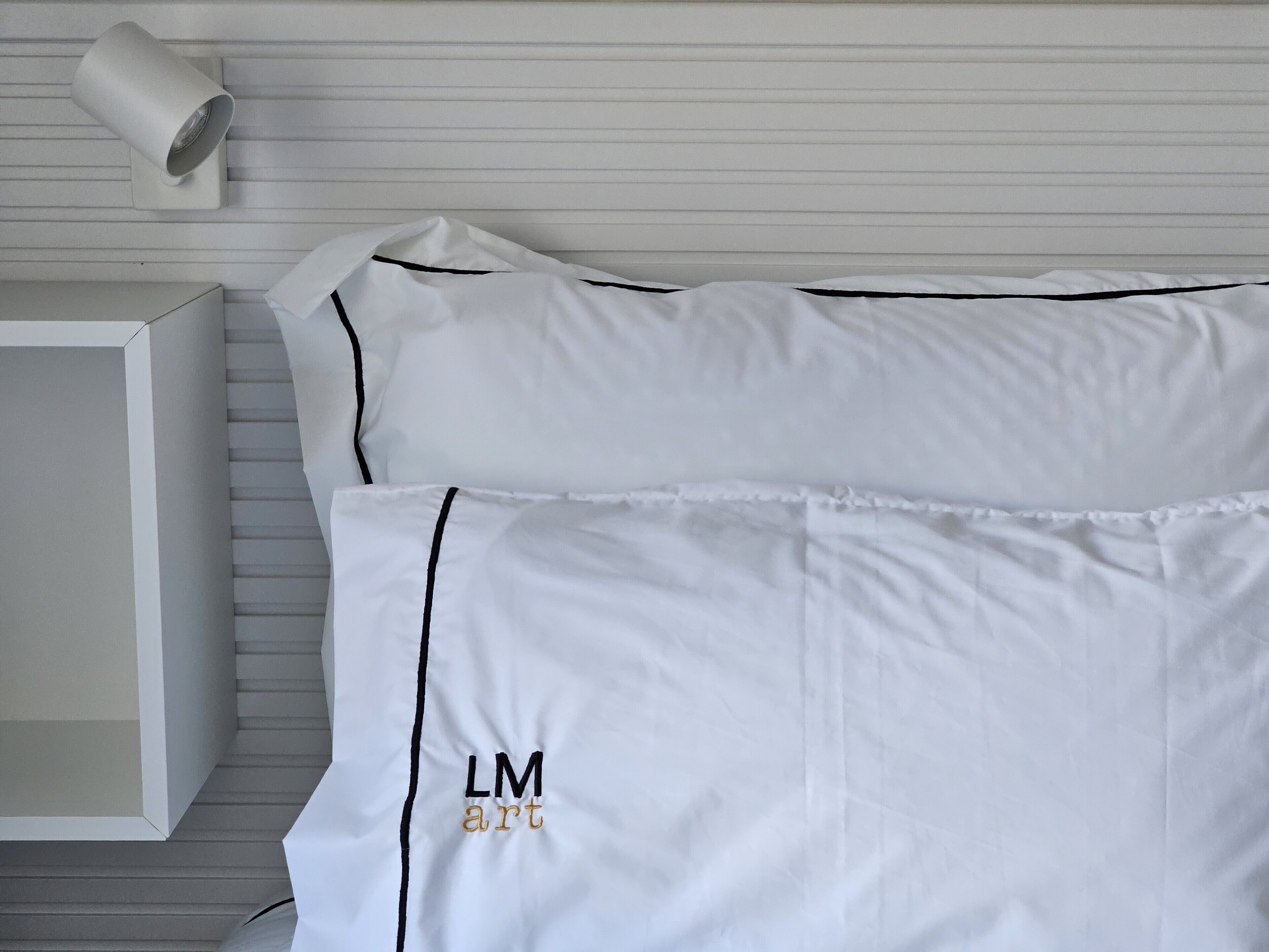 A neatly made bed with white linens and black piping, a wall-mounted light fixture, and a white bedside table.