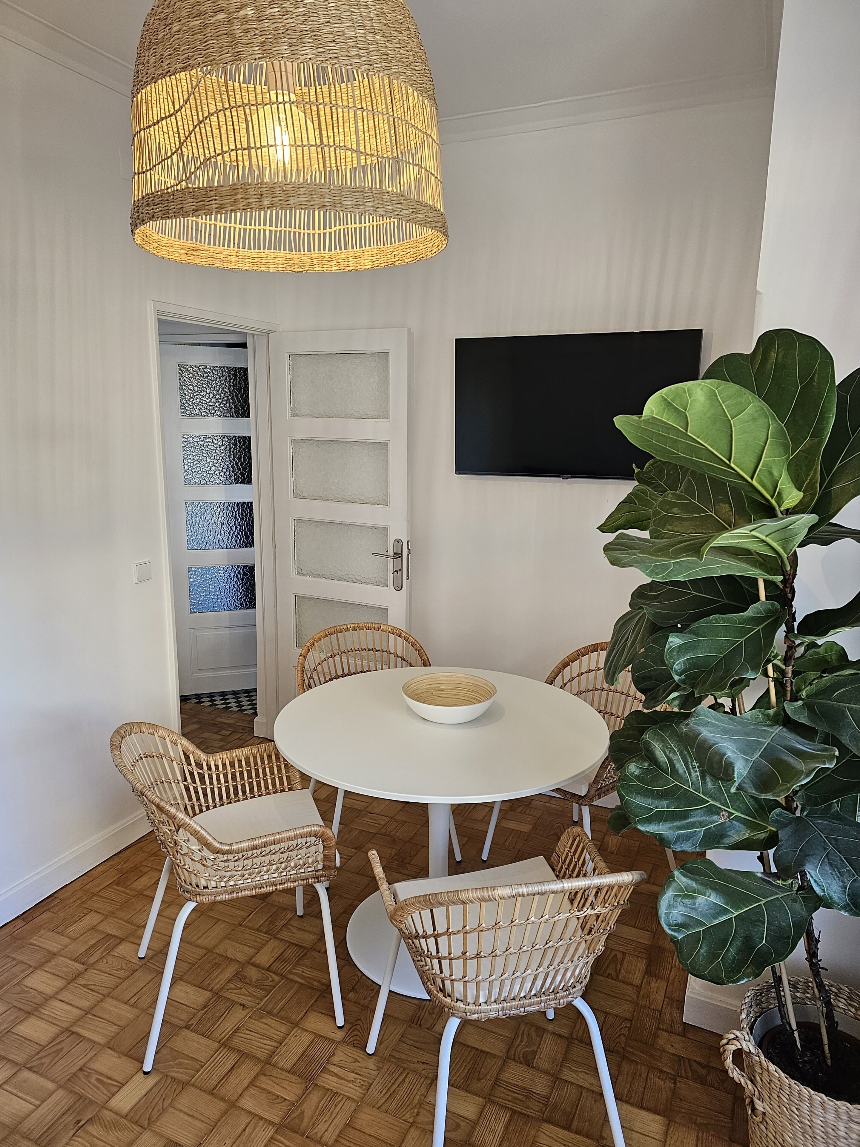 A dining area with a round table, wicker chairs, a woven pendant light, a large potted plant, and a television on the wall.