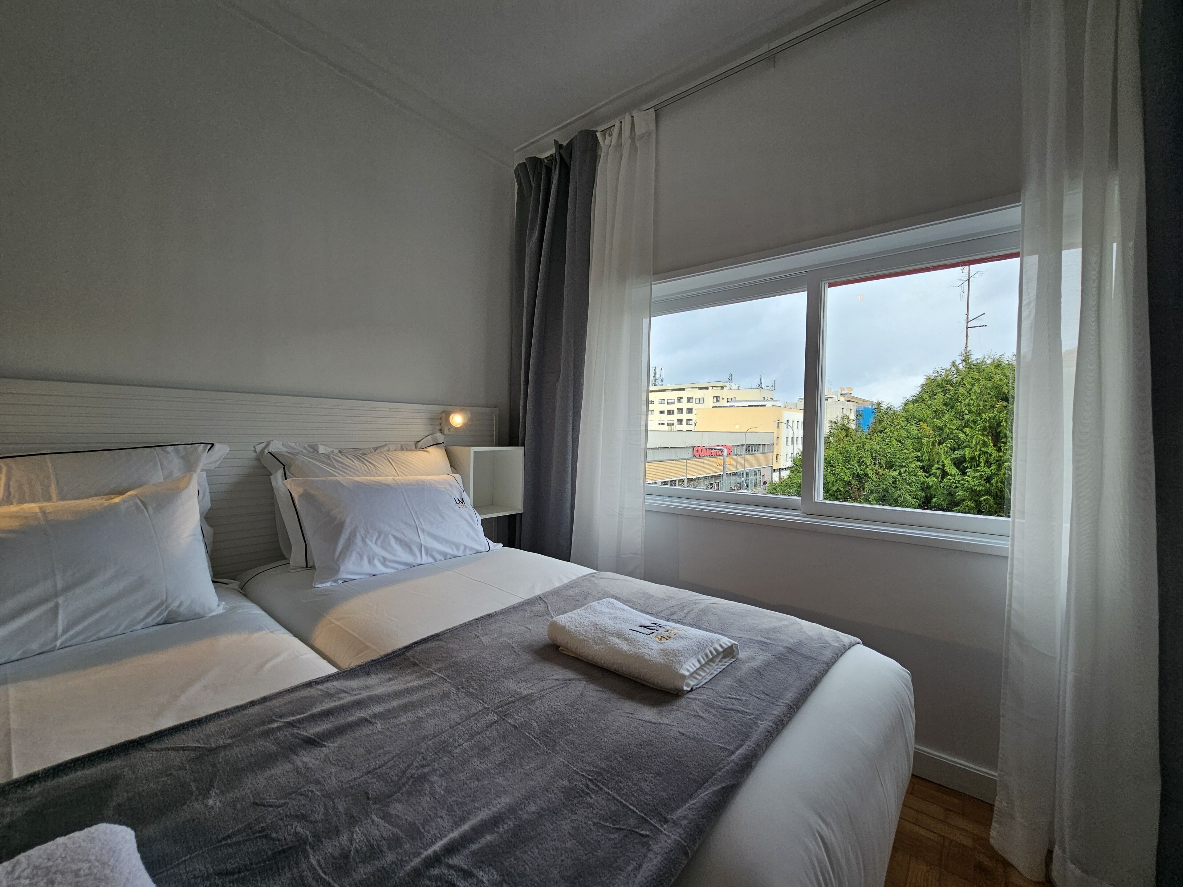 Une chambre d’hôtel avec un grand lit, une table de chevet avec une lampe et une fenêtre donnant sur des immeubles et des arbres.