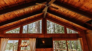 Une cabane avec un intérieur en bois, un ventilateur de plafond et de grandes fenêtres donnant sur la forêt.