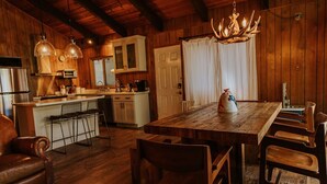 Un intérieur rustique en bois, avec une table à manger, des chaises, un canapé et un coin cuisine équipé d’appareils électroménagers et agrémenté d’un lustre.