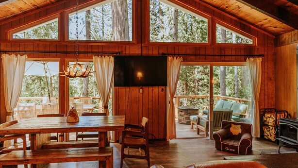 Une cabane en bois spacieuse, dotée de grandes fenêtres, d’un coin repas, d’une cheminée et d’un espace salon confortable.