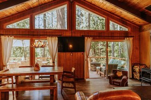 Une cabane en bois spacieuse, dotée de grandes fenêtres, d’un coin repas, d’une cheminée et d’un espace salon confortable.