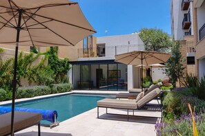 Un área de piscina moderna al aire libre con sillones y sombrillas, rodeada de exuberante vegetación y un cielo azul despejado.