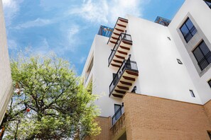 Un edificio de varios pisos con fachada blanca y base de ladrillo, que cuenta con balcones con barandas negras y un cielo azul despejado al fondo.