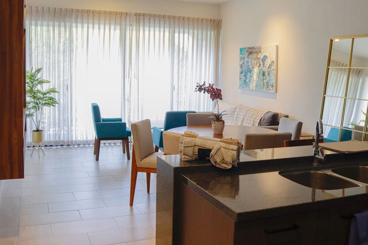 Una habitación de hotel moderna con área de comedor, sofá, mesa de centro, planta y un gran ventanal con cortinas.
