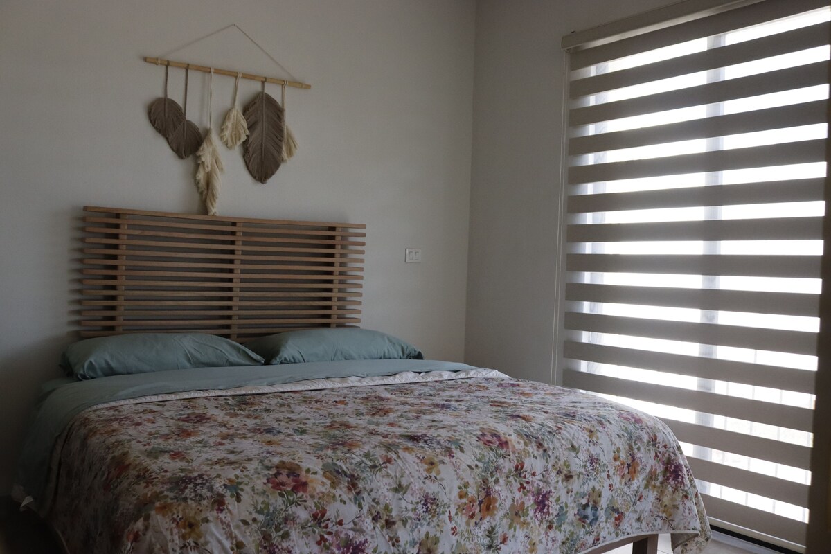 Un dormitorio con cabecera de madera con listones, una colcha con estampado floral y una ventana con persianas horizontales.