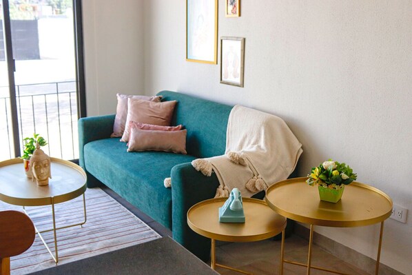 A teal sofa with a white throw blanket, two small round tables with gold frames, and a potted plant on one table.