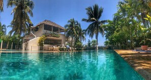 Une piscine à l’eau claire, entourée de palmiers et bordée d’un bâtiment à toit de chaume.