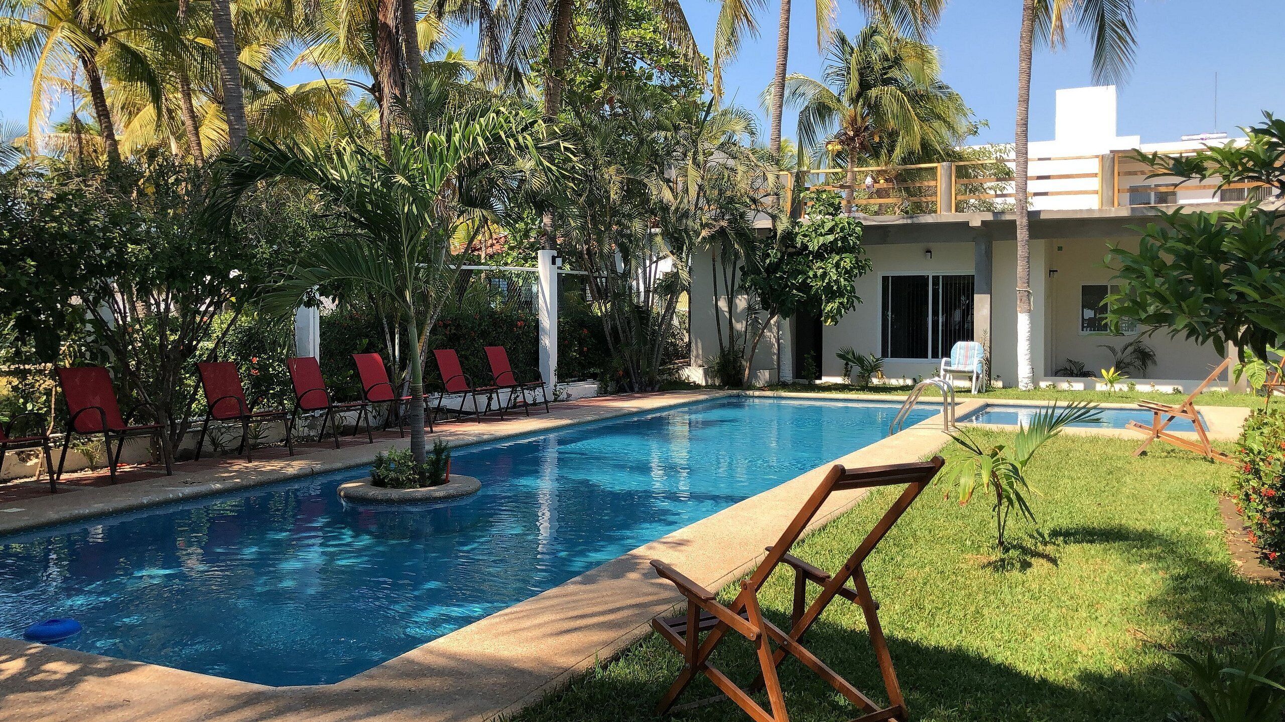 A swimming pool with red chairs, surrounded by palm trees and a building with a balcony.