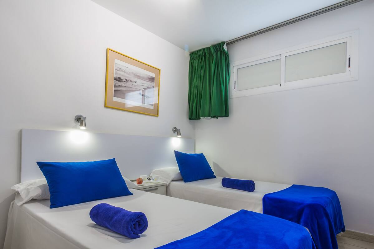 A hotel room with two beds, each with a blue bedspread and pillow, a white headboard, a framed picture on the wall, and a green curtain over the window.
