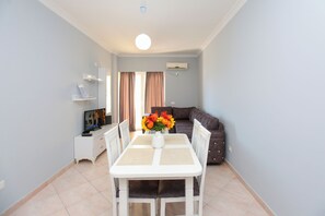 Ein Essbereich mit einem weißen Tisch, Stühlen und einer Blumenvase. Im Hintergrund befindet sich eine Couch und ein Fernseher.