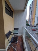 Un balcón con una pequeña mesa y sillas, vista a una calle con coches y un edificio alto al fondo.