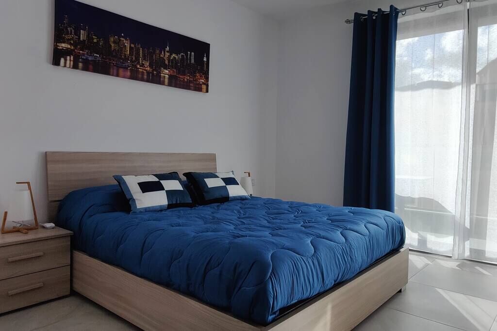 Un dormitorio con una cama grande, una mesita de noche, un cuadro de paisaje urbano y una ventana con cortinas.