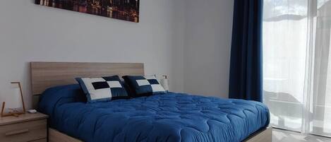 Un dormitorio con una cama grande, una mesita de noche, un cuadro de paisaje urbano y una ventana con cortinas.