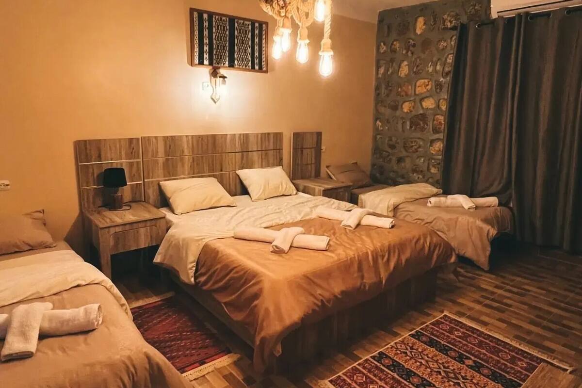 Habitación de hotel con tres camas, cabecera de madera, alfombra estampada y lámpara colgante.