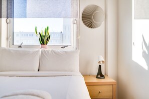 A bedroom with a bed, a bedside table with a lamp, a potted plant, and a wall-mounted mirror. - Cosy Battersea Flat (London)