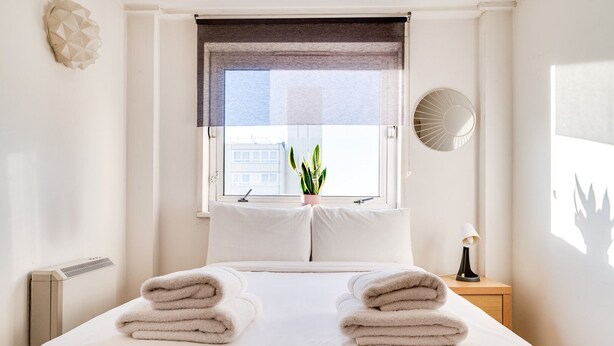 A neatly made bed with folded towels, a window with a view of buildings, a potted plant, and a wall-mounted fan. - Cosy Battersea Flat (London)