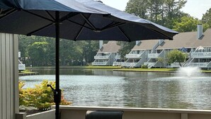 Vista al lago con una mesa y sillas en el patio, un parasol grande y un edificio con balcones al fondo.